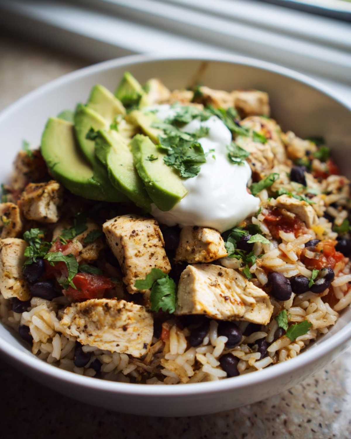A white bowl filled with seasoned chicken, rice, black beans, topped with sliced avocado, sour cream, and cilantro for a chicken burrito bowl.