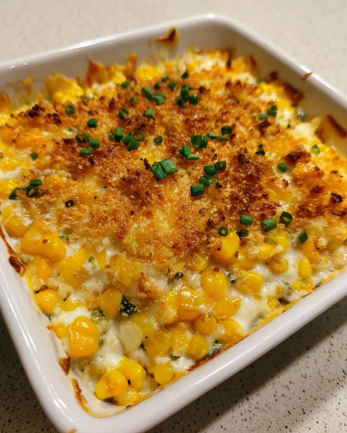 Close-up of a creamy, cheesy corn dip baked in a white dish with a golden breadcrumb topping and chives.