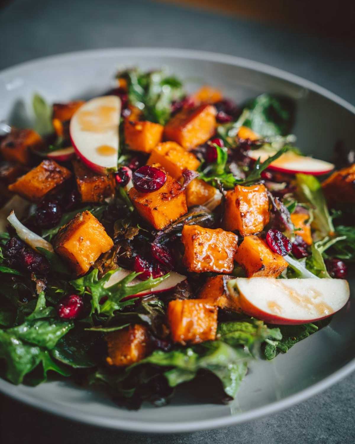 Close-up of an amazing fall harvest salad featuring roasted orange squash cubes, greens, apple slices, and cranberries.