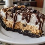 A decadent slice of layered ice cream pie featuring a dark crust, peanut butter filling, whipped topping, and chocolate drizzle.