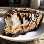 Close-up of a decadent slice of ice cream pie with peanut butter filling, whipped topping, and rich chocolate syrup drizzle.
