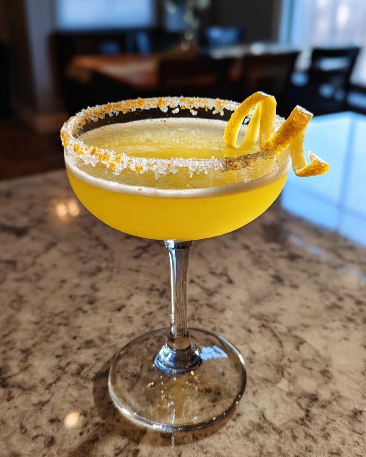 A bright yellow lemon drop cocktail served in a coupe glass with a sugar and zest rim and a lemon peel garnish.