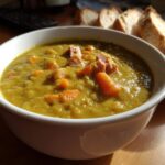 A close-up of a white bowl filled with thick, savory pea soup, topped with chunks of ham and carrots.