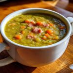 Close-up of a thick, green pea soup in a white mug, topped with chunks of ham and diced carrots.