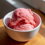 Two scoops of bright pink, textured strawberry ice cream served in a small white bowl.
