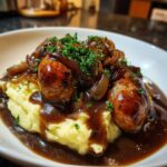 A close-up of delicious bangers and mash, featuring sausages, creamy mashed potatoes, rich gravy, and caramelized onions.
