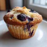 Close-up of a single, fluffy blueberry cottage cheese muffin topped with coarse sugar, resting on a white plate.