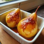 Two glistening baked pears covered in a rich caramel glaze sitting in a white baking dish.