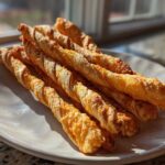 A pile of golden, flaky, twisted cheese straws recipe resting on a light-colored plate near a window.