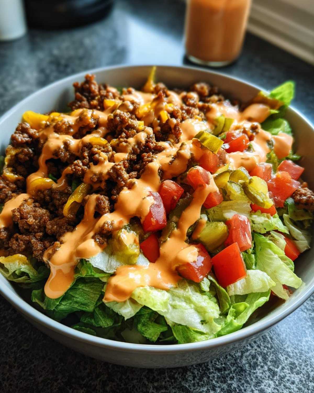 A close-up view of a healthy cheeseburger bowl featuring ground beef, lettuce, tomatoes, pickles, and a special sauce drizzle.