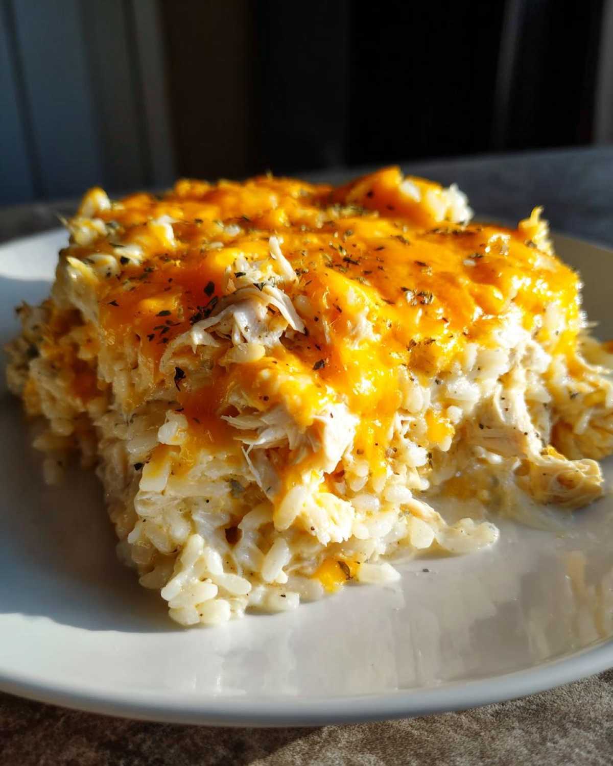 A square serving of cheesy chicken and rice topped with melted cheddar and herbs on a white plate.