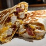 Close-up of a grilled chicken bacon ranch wrap cut in half, showing chunks of chicken, bacon, and melted cheese.