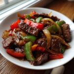 A close-up shot of glossy, saucy chinese pepper steak mixed with vibrant green and red bell peppers and onions in a white bowl.