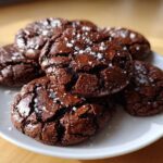 A stack of fudgy chocolate brownie cookies topped with flaky sea salt on a white plate.