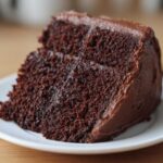 Close-up of a moist, dark slice of chocolate cake recipe covered in thick chocolate frosting on a white plate.