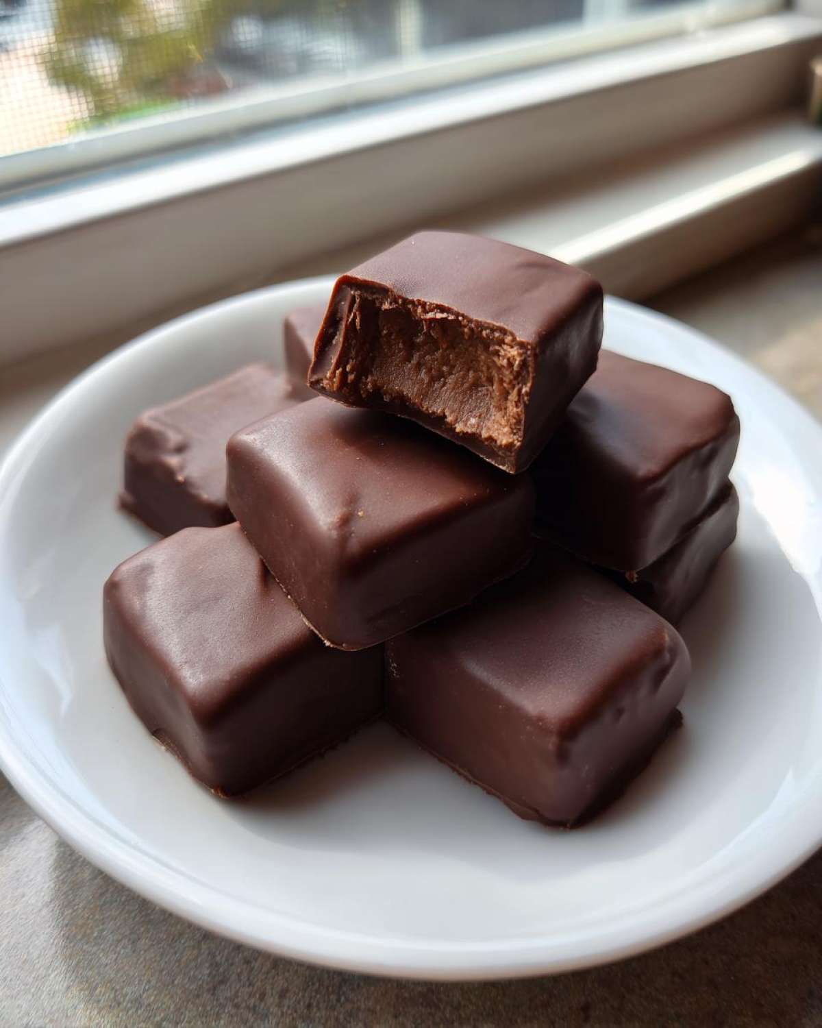 A stack of chocolate-covered cool whip candy pieces on a white plate, one piece has a bite taken out.
