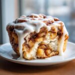 A close-up of a single serving of cinnamon roll apple pie, showing the soft dough, apple filling, and thick white icing.