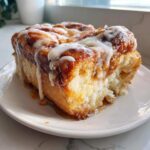 A close-up, appetizing shot of a thick slice of cinnamon roll french toast drizzled heavily with white icing.