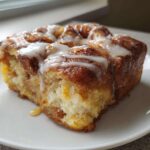 A close-up of a square serving of cinnamon roll french toast, drizzled with white icing, on a white plate.