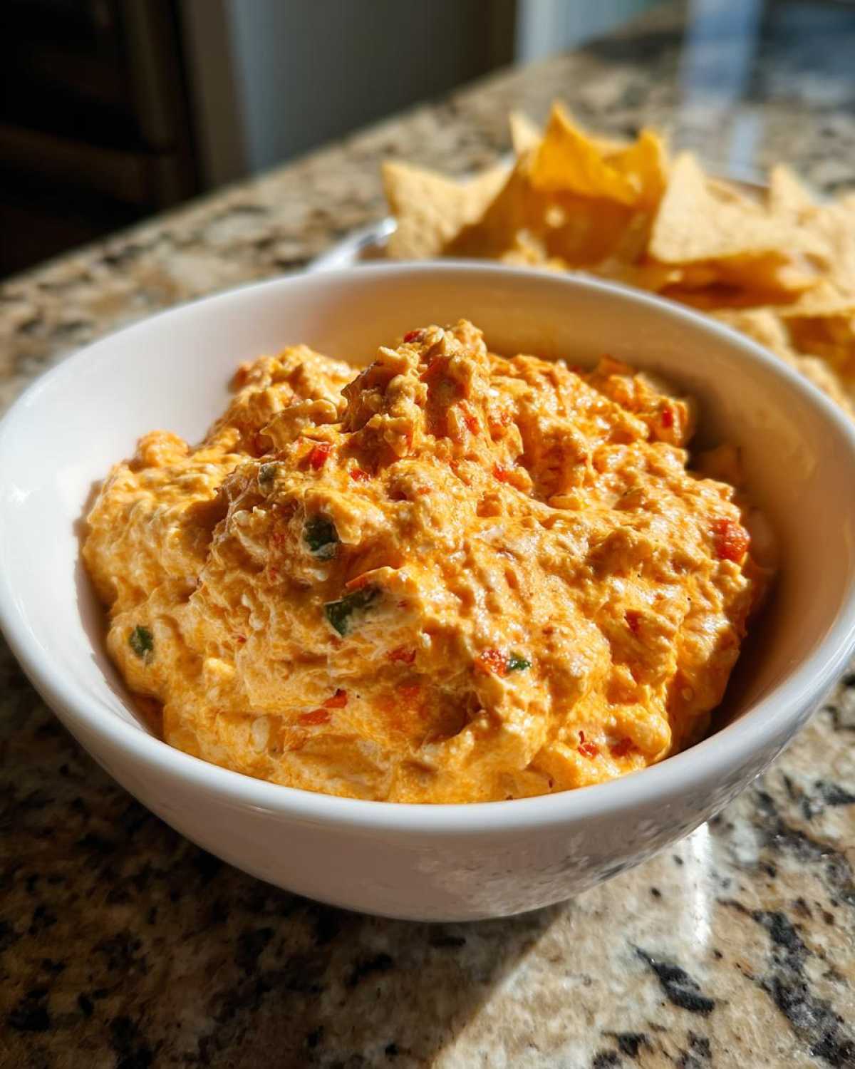 Close-up of a thick, orange boat dip mixed with peppers, served in a white bowl with tortilla chips blurred in the background.