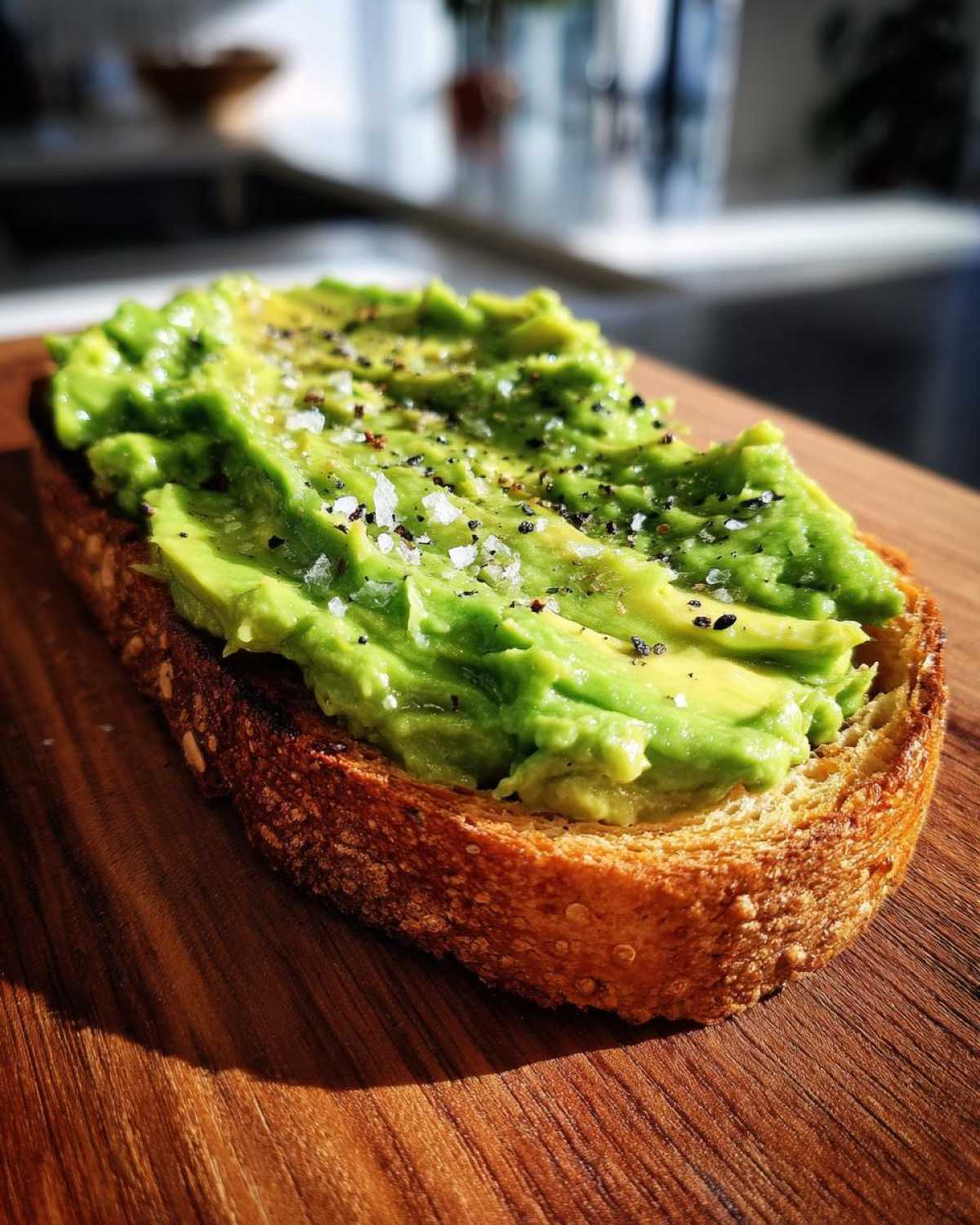 Close-up of creamy avocado toast topped with flaky salt and cracked black pepper on a wooden board.