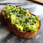 Close-up of creamy avocado toast topped generously with flaky salt, cracked black pepper, and fresh herbs.