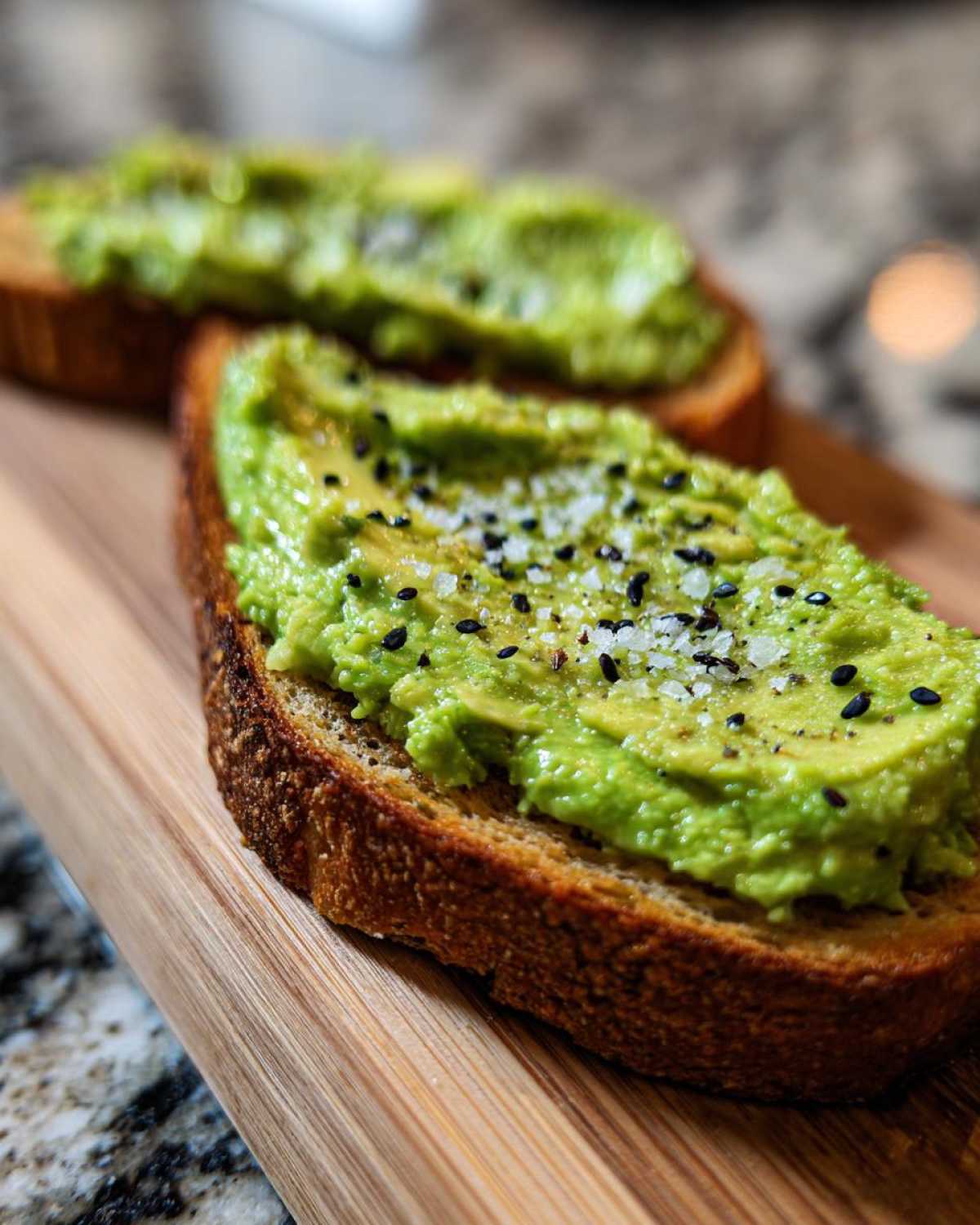 Close-up of creamy avocado toast topped with flaky salt and black sesame seeds on a wooden board.