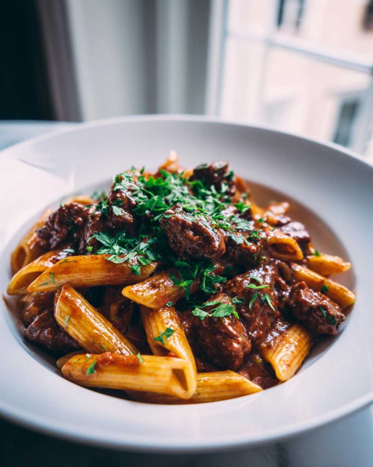 A close-up of a white bowl filled with creamy beef pasta (penne) topped generously with fresh chopped parsley.