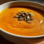 Close-up of a bowl of vibrant orange, creamy butternut squash soup garnished with toasted pumpkin seeds.