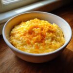 A close-up of a white bowl filled with creamy, yellow cheese grits topped with freshly shredded cheddar cheese.