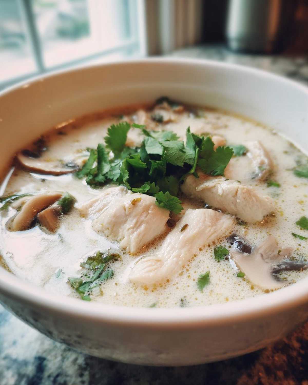 A close-up of a white bowl filled with creamy chicken coconut soup, featuring sliced chicken breast, mushrooms, and fresh cilantro.