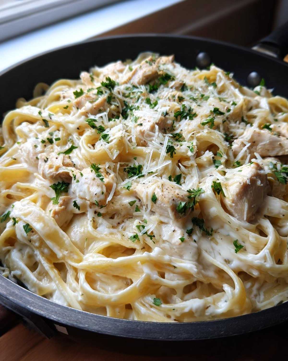 Close-up of creamy chicken fettuccine alfredo pasta topped with grated parmesan and fresh parsley in a black skillet.