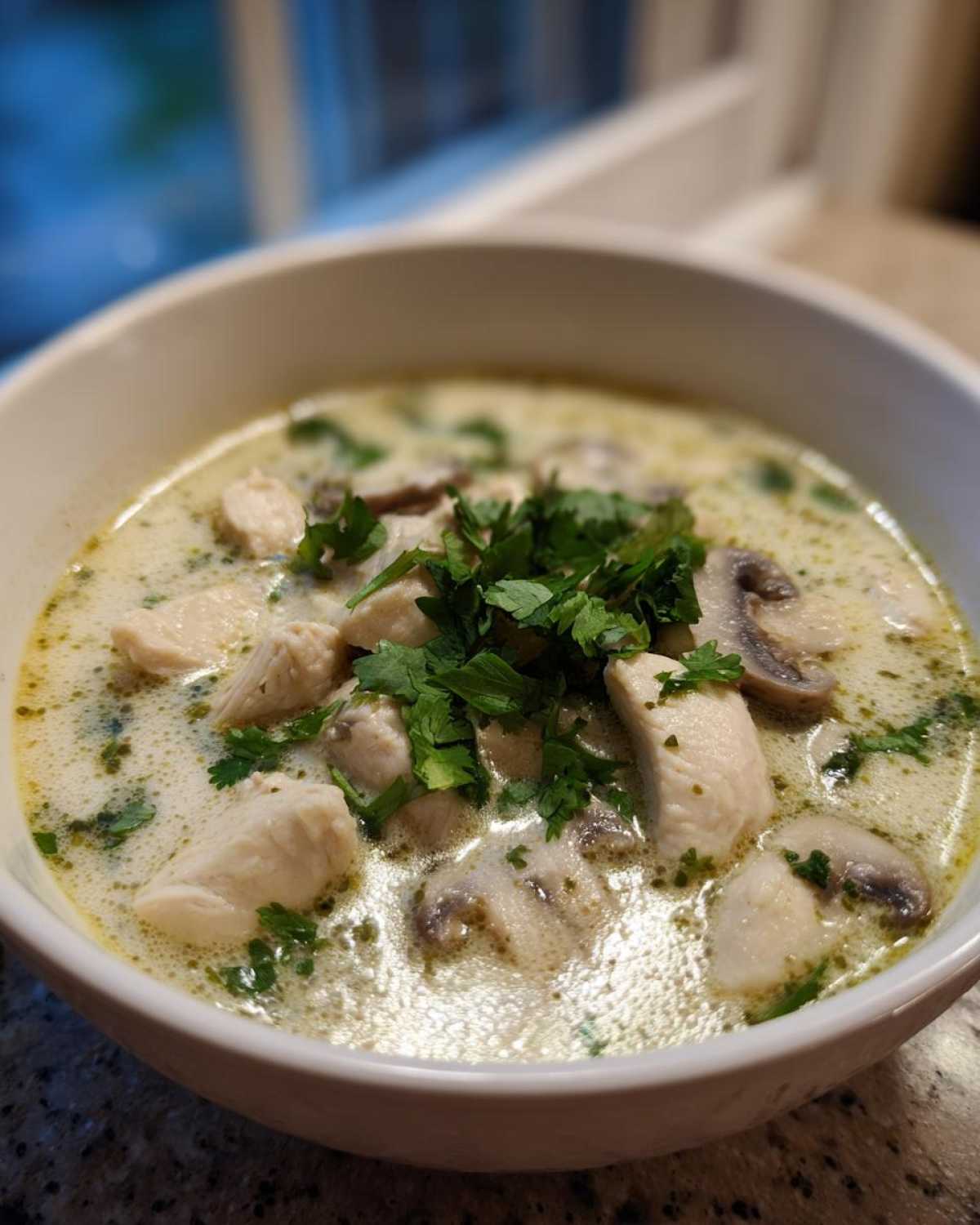 Close-up of a white bowl filled with creamy coconut soup, featuring chunks of chicken, sliced mushrooms, and fresh parsley garnish.