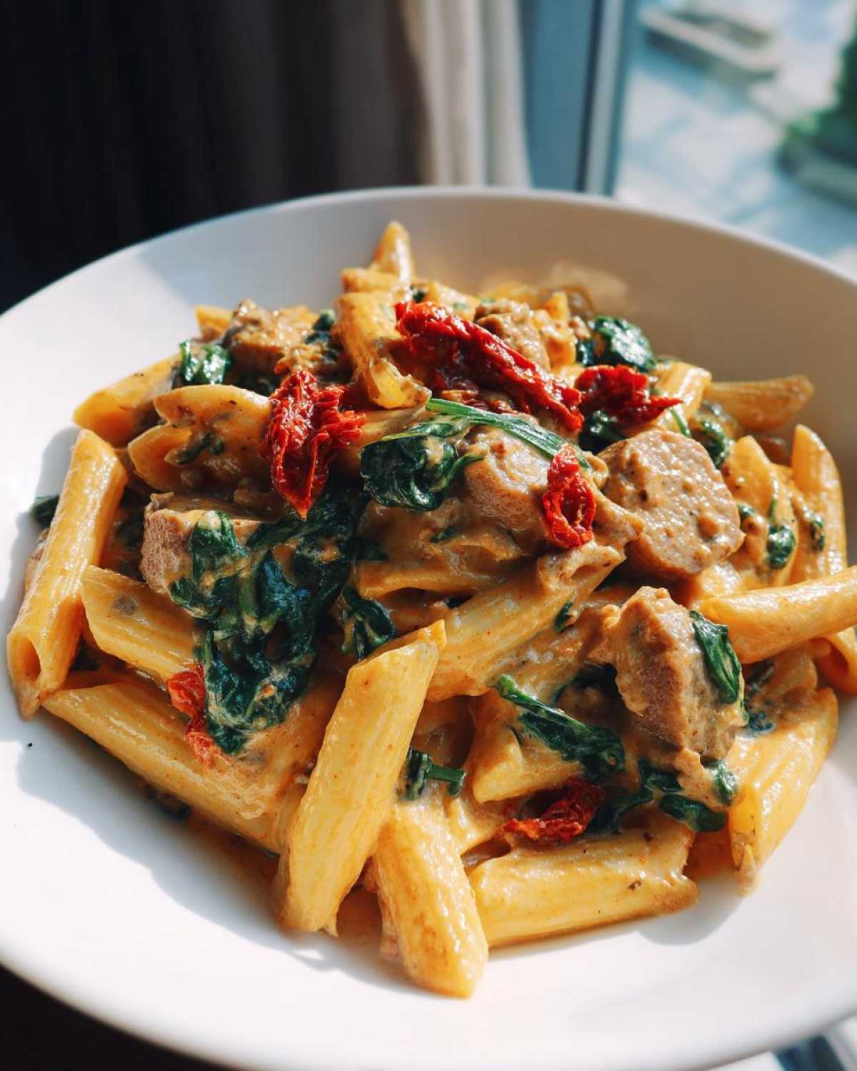 Close-up of creamy chicken sausage pasta featuring penne, spinach, and sun-dried tomatoes in a white bowl.