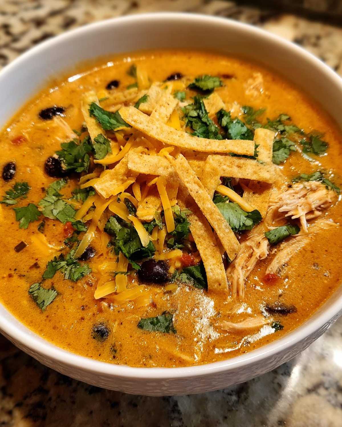 Close-up of a bowl of creamy chicken tortilla soup topped with shredded cheese, cilantro, and tortilla strips.