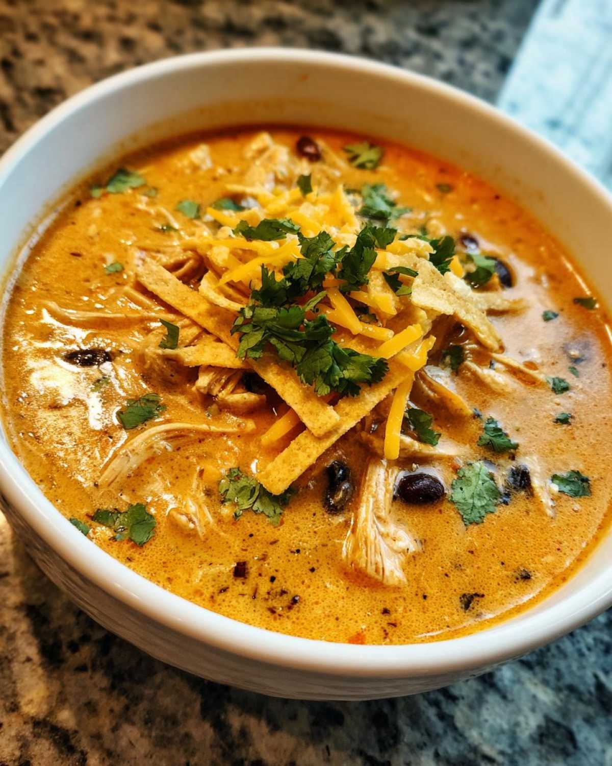 Close-up of a white bowl filled with creamy chicken tortilla soup, topped with shredded cheese, cilantro, and tortilla strips.