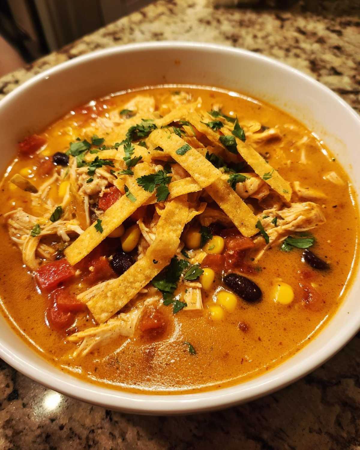 A close-up of a bowl of creamy chicken tortilla soup topped with tortilla strips, corn, beans, and cilantro.