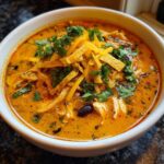 Close-up of a bowl of creamy chicken tortilla soup topped with shredded cheese, tortilla strips, and cilantro.