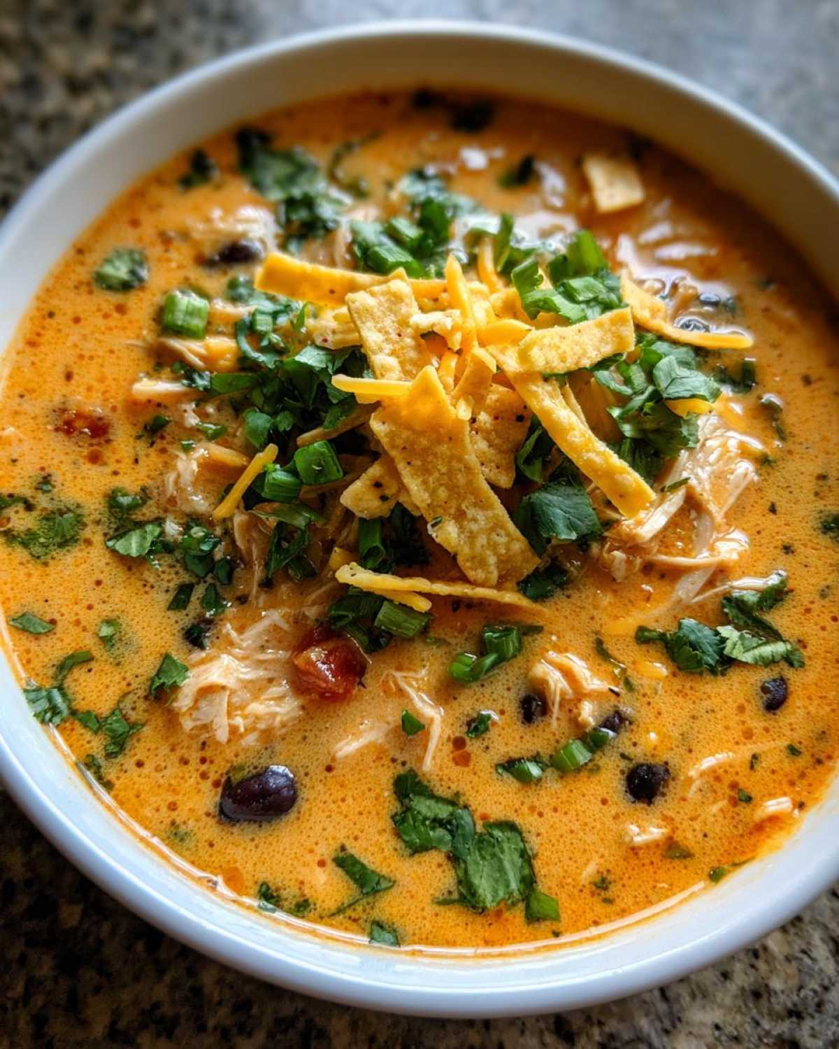 Close-up of a bowl of creamy chicken tortilla soup topped with shredded chicken, black beans, cilantro, green onions, and tortilla strips.