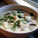 Close-up of a white bowl filled with creamy coconut soup, featuring chunks of chicken, mushrooms, and fresh cilantro.