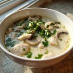 Close-up of a white bowl filled with creamy coconut soup, featuring chunks of chicken, sliced mushrooms, and fresh cilantro.
