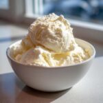 A white bowl filled with several scoops of creamy, pale yellow cottage cheese ice cream, brightly lit by a window.