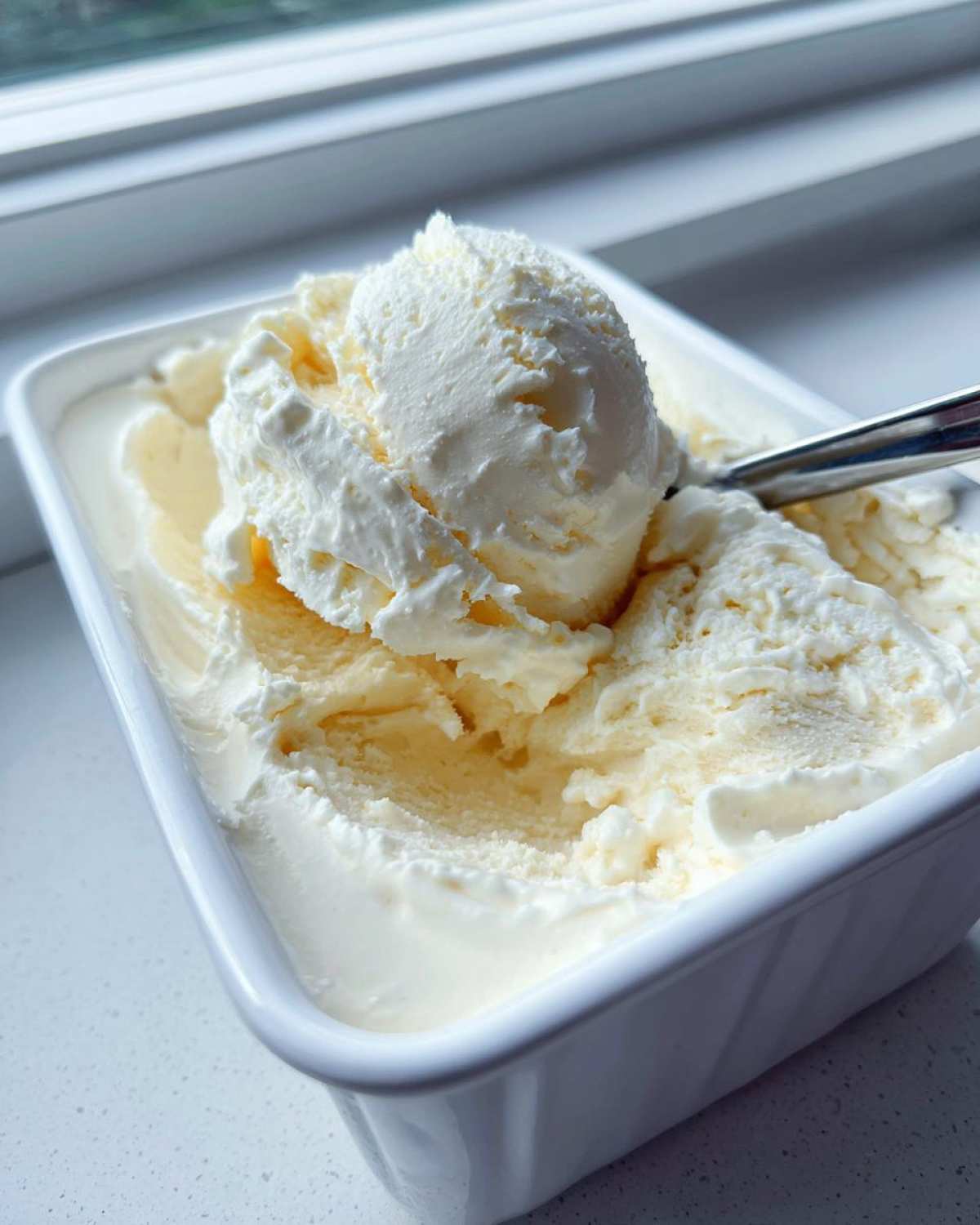 A scoop of creamy, vanilla-colored cottage cheese ice cream being lifted from a white container with a spoon.