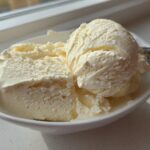 Close-up of creamy, pale yellow cottage cheese ice cream scoops in a white bowl near a window.