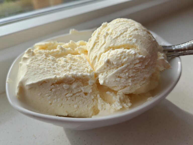 Close-up of creamy, pale yellow cottage cheese ice cream scoops in a white bowl near a window.