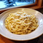 A white bowl filled with creamy spaghetti topped generously with grated parmesan cheese, suggesting a delicious garlic parmesan pasta.