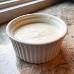 Close-up of creamy white garlic sauce served in a small, white, fluted ramekin on a granite countertop.