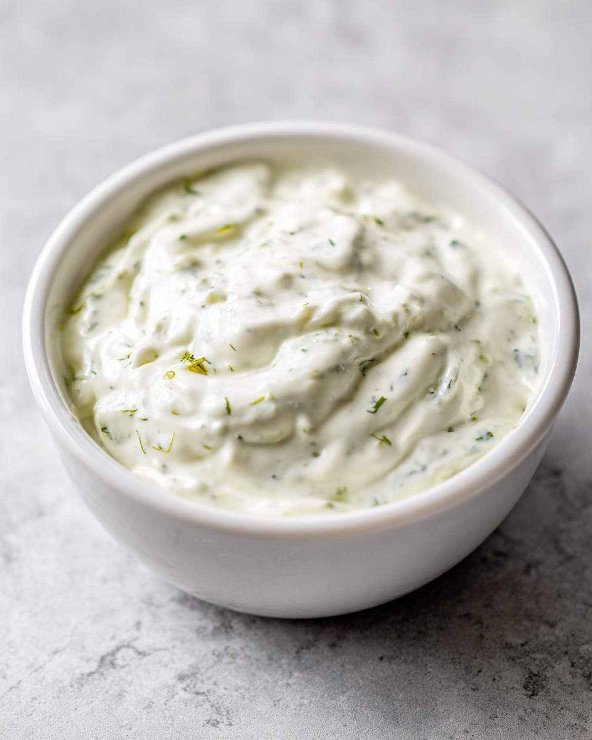 Close-up of creamy, white homemade tartar sauce with visible green herbs in a small white bowl.