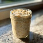 A glass jar filled with creamy overnight oats, including visible oats and chia seeds, sitting on a granite counter in bright sunlight.