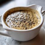 Close-up of rich, creamy peppercorn sauce generously topped with cracked black peppercorns in a small white serving dish.